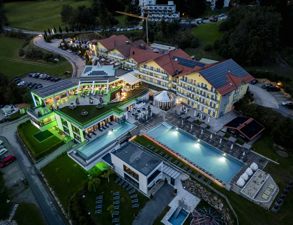 Die Adresse für Wellness in Bayern: Ihr Hotel in St. Englmar Luftaufnahme eines beleuchteten Hotels mit Außenpool und moderner Architektur bei Dämmerung